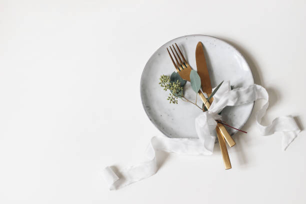 réglage de table festif. coutellerie dorée, branche d'eucalyptus de baies, plaque de porcelaine et ruban de soie isolé sur le fond blanc de table. concept méditerranéen de menu de mariage ou de restaurant. flat lay, vue du haut. - plate silverware table white photos et images de collection