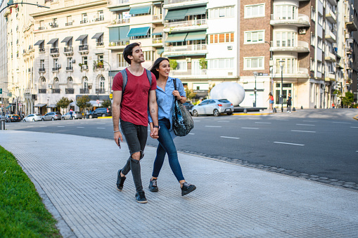 Relaxed Hispanic tourists in their 20s holding hands and walking in downtown Buenos Aires on sunny weekend city break.