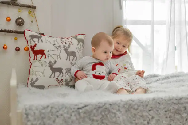 Photo of Brother and sister in Christmas pajamas are sitting on the bed.