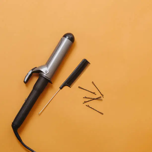 Hairdresser tools: curling iron, rat tail comb and bobby pins over orange background. Top view. Necessary things for styling hair, including: curling, straightening, separating, sectioning hair.