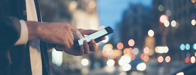 Closeup image of male hands with smartphone at night on city street, searching internet or social networks, hipster man typing an sms or message on chat, bokeh lights