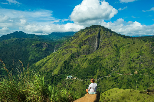 Caucasian tourist woman sitting on stone on Mini Adams peak looking on beautiful valley and green mountains of Sri Lanka