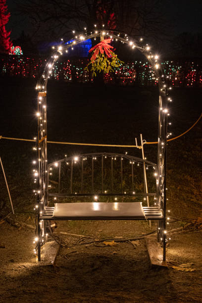 Place for kisses on empty bench with arch and mistletoe branch in night lights. Shiny lights garlands and red ribbon on mistletoe hanging in arch in darkness. Christmas tradition. Love concept stock photo