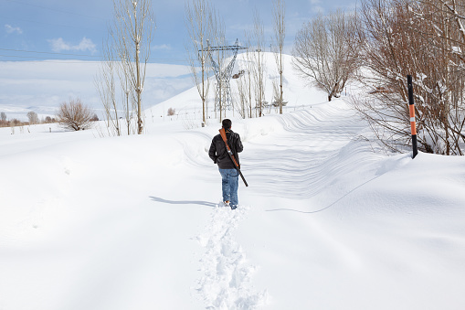 Rear view of man with rifle walking on snow in countryside. Bare trees are seen around. Shot in wintertime during day with a full frame DSLR camera.