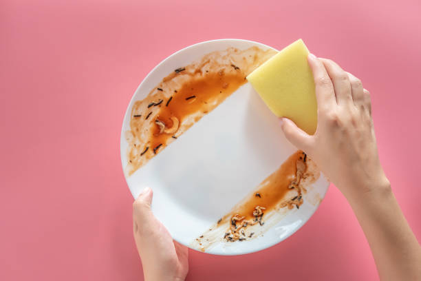 mujer usando esponja de limpieza amarilla para limpiar y lavar las manchas de alimentos y la suciedad en el plato blanco después de comer comida aislada sobre fondo rosado. limpieza, salud y saneamiento en el hogar concepto - cleaning sponge fotografías e imágenes de stock