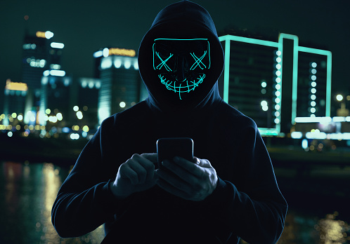 Portrait of an anonymous man in a black hoodie and neon mask hacking into a smartphone. Bright city background