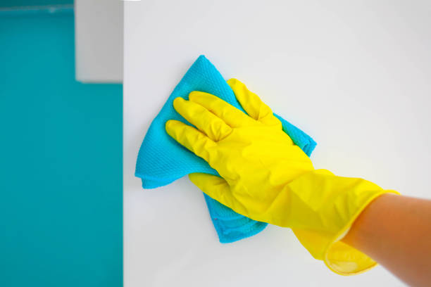 A female hand in yellow rubber gloves wipes a modern white kitchen with a microfiber cloth.