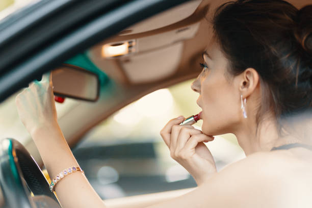 Young elegant woman looking in the car view mirror while applying makeup, lipstick on the lips