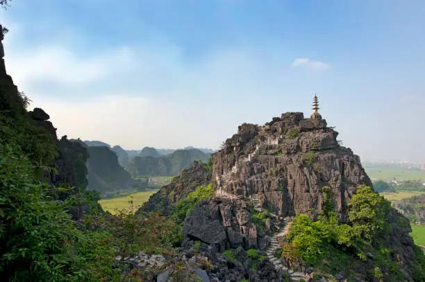 Hang Mua temple stands out from the lush greens in the flat paddy fields surrounding the dramatic moody limestone mountains of Tam Coc, Ninh Binh, in the rural scenery of woodland covered hills, from above at Hang Mua high viewpoint as the rice crop shoots ripen during the harvest in Tam Cốc-Bích Động, Ninh Binh Province, North Vietnam