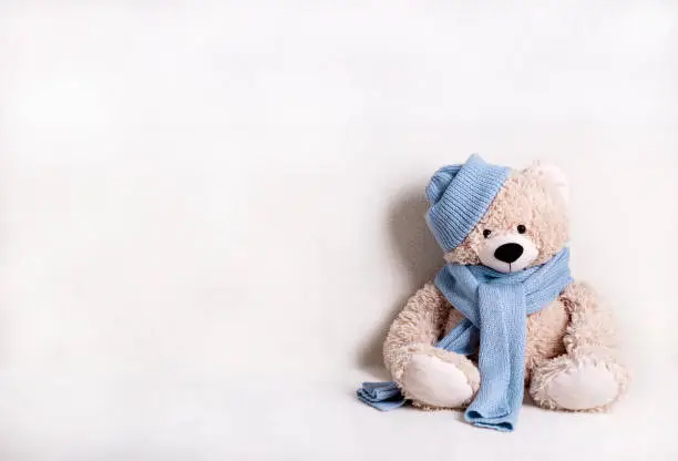 Photo of Children's toy teddy bear is sitting on a white sofa in a knitted scarf and hat in blue.