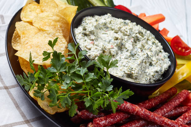 Creamy spinach dip in a bowl with sausages, carrots, cucumber, sweet pepper sticks, potato chips and fresh parsley on a plate close-up of Creamy spinach dip in a bowl with sausages, carrots, cucumber, sweet pepper sticks, potato chips and fresh parsley on a plate on a white table,  view from above, dipping stock pictures, royalty-free photos & images