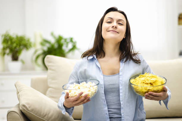 Happy and pleased woman holds deep plates with pop corn and chips. She is ready to film watching. Concept leisure; junk food. Film watching with snacks. Copy space. deep pick stock pictures, royalty-free photos & images