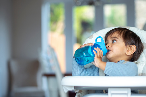 A front-view shot of a young boy drinking water from a reusable water bottle at home, he is wearing casual clothing and sitting in a high chair.