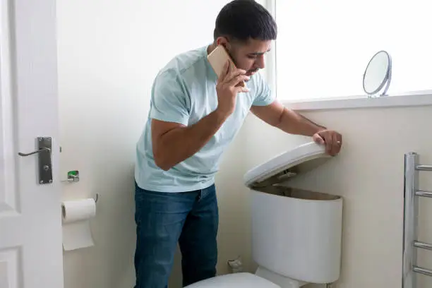A side-view shot of a man standing in his bathroom looking at his toilet, there is a problem, he is on the phone trying to call a plumber.