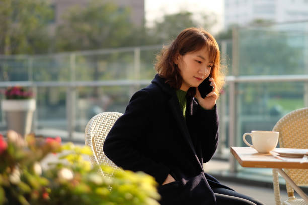 une belle jeune femme asiatique s'asseyant à l'extérieur sous le soleil, faisant l'appel de téléphone. - autumn table setting flower photos et images de collection