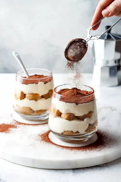 Traditional Italian dessert tiramisu in a glass. Female hand sprinkling cocoa powder over tiramisu. Woman pours cocoa powder