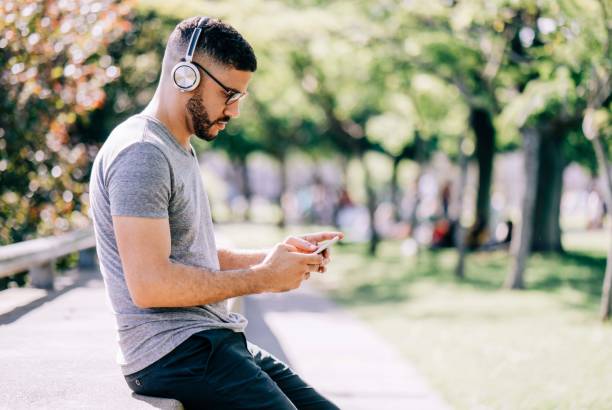 Side view of man using mobile phone while sitting in park. Male is listening music through wireless headphones. He is spending leisure time.