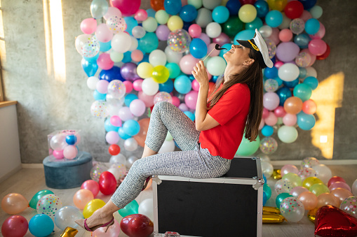 Attractive young woman sitting on metal box in studio with colorful balloons on wall, wearing sailor hat