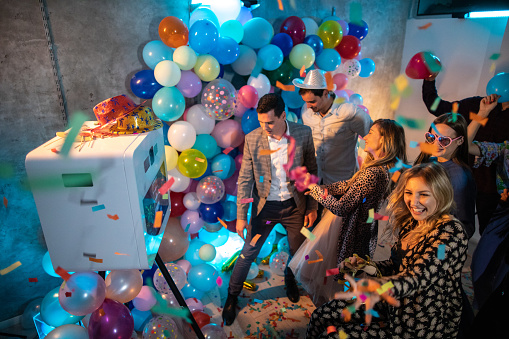Young woman popping a confetti blaster while her friends taking photo on photo booth machine during party