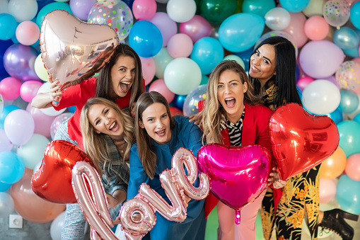 Five beautiful women standing in front of wall with balloons and holding helium balloons in hand, looking at camera and smiling