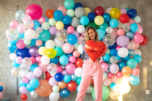 Portrait of beautiful young woman standing in front of wall with large group of balloons on it, holding hearth shape helium balloon, looking at camera and smiling