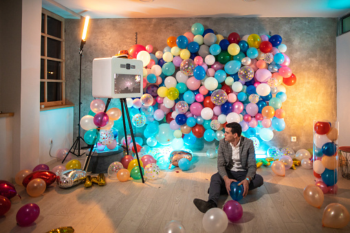 Young man sitting on floor after party, holding balloon and looking at photo booth