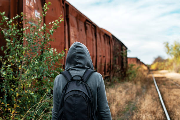 Homeless woman with backpack getting away from it all, rear view of female walking among abandoned train wagons and obsolete railroad track