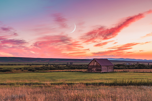 A beautiful sunset in a rural Montana scene. A simple barn sits in a field as the sun sets while the crescent moon rises above.