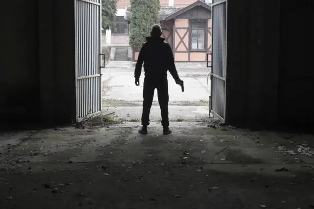 Photo of Silhouette of man standing at large entrance of spooky, abandoned building holding handgun replica in his hand