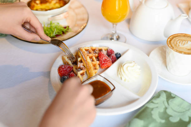 petit déjeuner ou brunch du matin au restaurant. table avec des boissons et de la nourriture. les mains des femmes coupent les gaufres viennoises avec un couteau et une fourchette. orientation sélective - snack coffee instant coffee cappuccino photos et images de collection