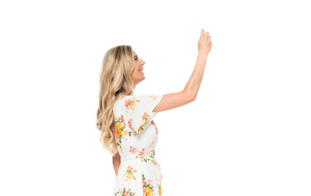 side view / profile view / waist up of 20-29 years old adult blond hair / long hair caucasian young women standing wearing dress / long dress who is smiling / happy / cheerful who is taking a selfie / photographing and holding mobile phone - 20 25 years profile female young adult imagens e fotografias de stock