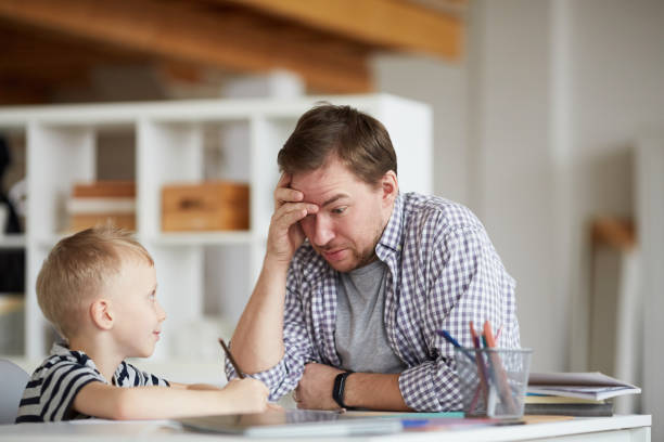 istock Confused young father puzzled with sons home task solution sitting at table and holding head in hand while his son looking at him in anticipation of answer 1192951145