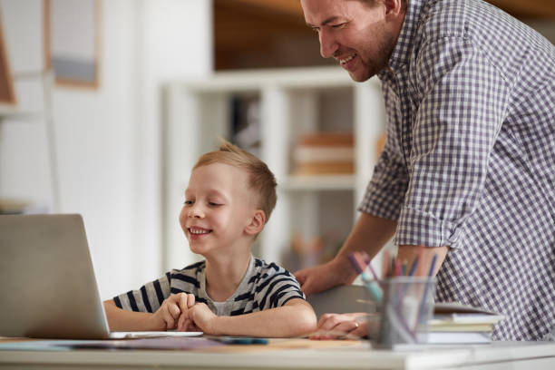 istock Cheerful cute boy sitting at table and looking at laptop screen with father while they using modern computer app for education 1192951177
