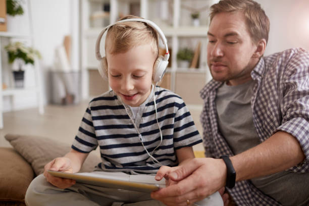istock Positive cute blond-haired boy in wired headphones using learning app on tablet together with father at home 1192951228