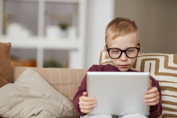 istock Serious curious little boy in glasses sitting on sofa and using digital tablet while reading online book 1192951306
