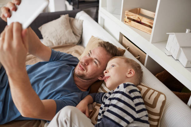 istock Smiling handsome young father with stubble lying on bed and using portable device while making self-portrait with son sticking tongue out 1192951323
