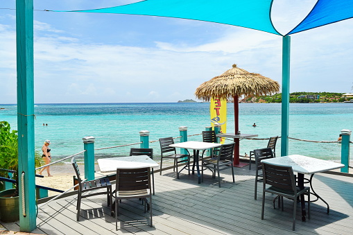 Outside restaurant on island of St. Thomas, beach side.  Blue tarpon sun shield covers over restaurant tables and chairs on stone patio with aqua blue Caribbean and tiki umbrella.