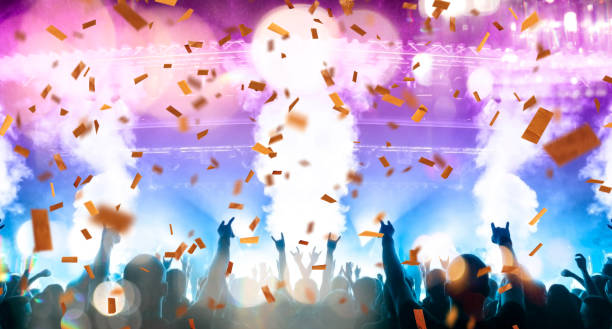 Concert stage, people are visible waving and clapping, silhouettes are visible