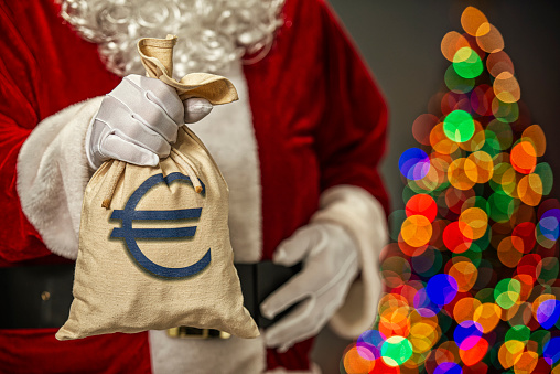 Santa Claus holding a canvas Euro money bag with blue € symbol and in the background the bokeh of multi-colored Christmas tree lights