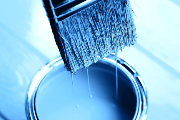 Blue paint is dripping from brush into open can on freshly painted wooden background. Renovation concept. Macro.