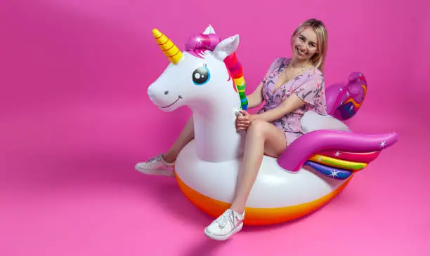 Photo of A beautiful blonde girl in a sexy sundress with slim legs in white sneakers sits on an inflatable multi-colored unicorn on a pink isolated background. Summer vacation at the beach and travel.