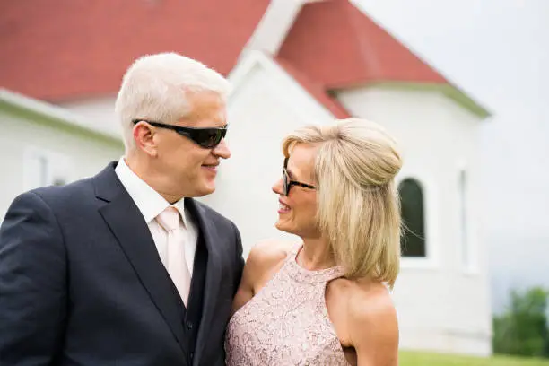 Blond woman wearing a pale pink lace long dress stands with her husband wearing a dark suit outdoors before their daughter's wedding, Minnesota, USA
