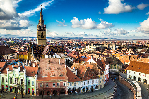 Color image depicting the ancient, medieval skyline and anglo-Saxon architecture of Sibiu, a city in the Transylvania region of Romania. People are wandering and exploring Piata Mica, and we can see the so-called Bridge of Lies in the distance. Room for copy space.