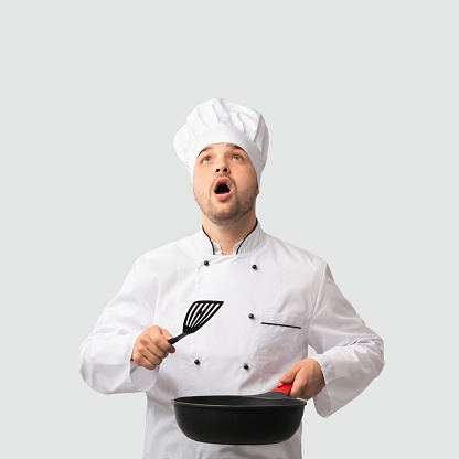 Cooking Food. Surprised Chef Man Holding Pan And Spatula Looking Up Standing Over White Background. Studio Shot