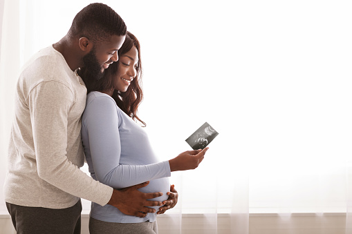 Emotional black couple holding sonogram picture over white background at home, expecting baby, free space
