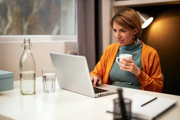 Smiling dedicated hardworking Caucasian pregnant businesswoman sitting in office, using laptop and drinking tea.