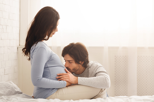 Young future dad tenderly kissing belly of his pregnant wife while spending time together at home, copy space
