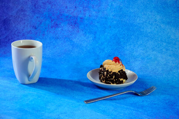 A high white ceramic mug with tea, next to the same saucer with cream and cherry cake, next to a spoon, the whole composition on a blue abstract background.