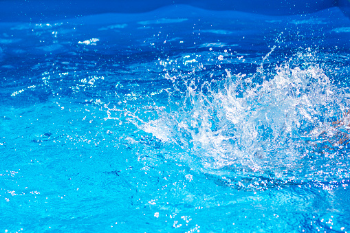Splashes of clear blue fresh water in pool, air bubbles, water drops, sea wave on blue background with sunny reflections.