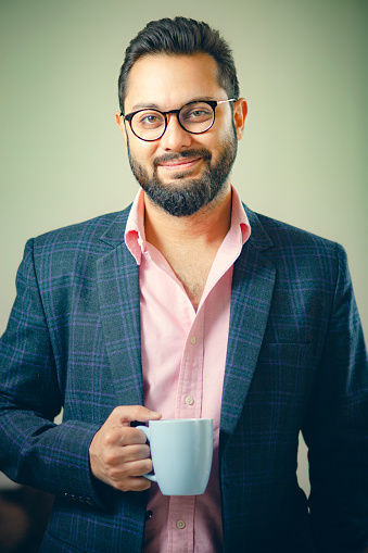 An Asian/Indian confident and handsome businessman holds a coffee mug. He wears a formal dress and looks at the camera with a smile.